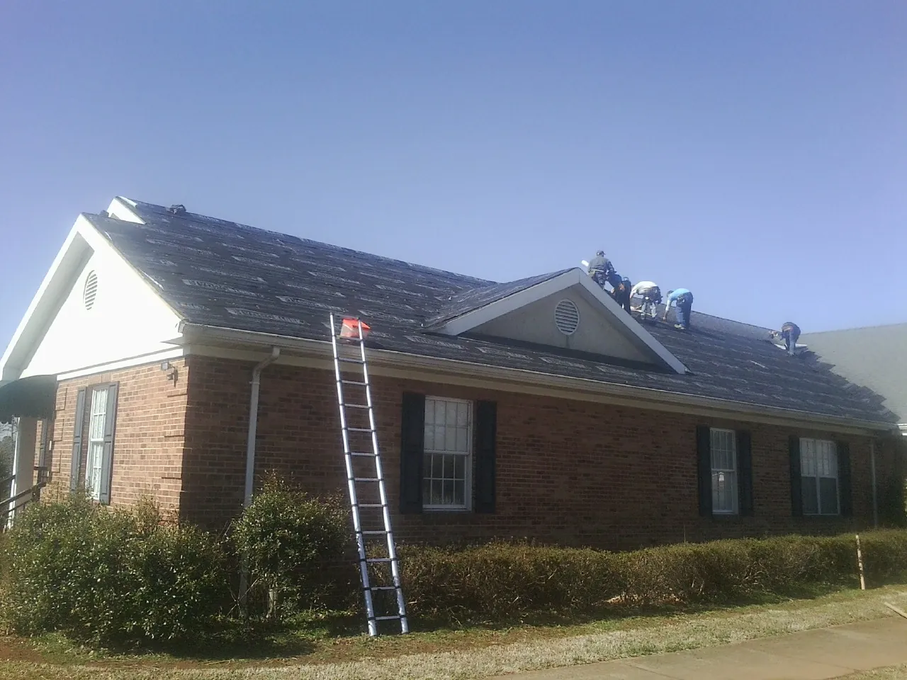 Crew tearing shingles off a brick ranch roof with a ladder set — Emergency Roof Repair work by our crew in Halbur