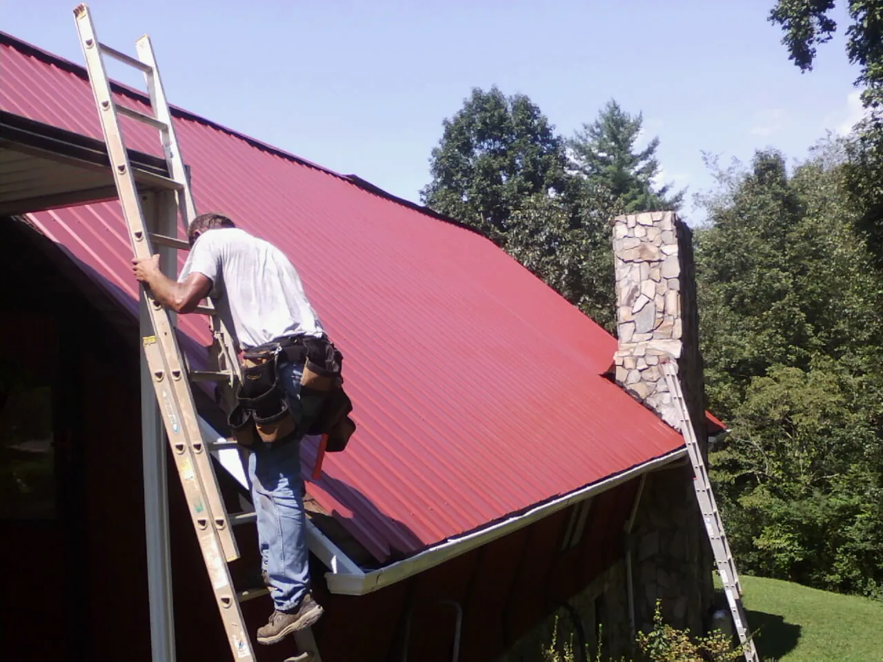 Roofer on a ladder beside a red metal roof and stone chimney — Soffit & Fascia Repair by Roofing Brothers in Halbur